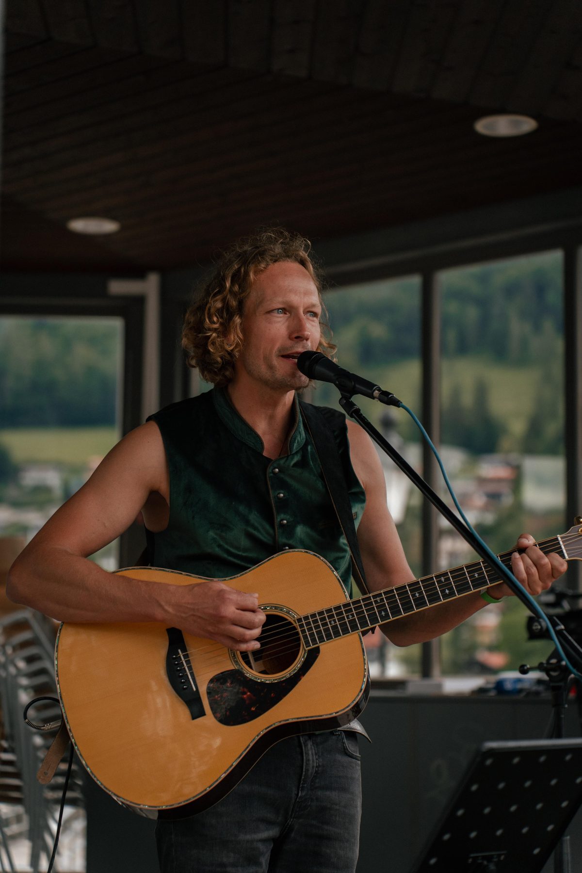 Tobi Sonic singt ins Mikrofon und spielt Akustikgitarre, im Hintergrund Berge hinter großen Fenstern